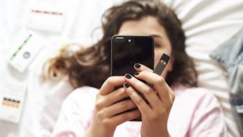 A teenage girl looks at her phone while holding a JUUL vaping device. Learn more about vaping and how it impacts youth.