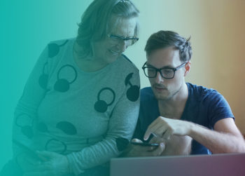 Two people wearing glasses look down at a digital device, one wearing a gray sweater with headphone patterns and the other in a navy v-neck shirt, bathed in turquoise and amber lighting.