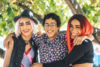 Portrait of three LGBTQ friends outdoors, smiling, in front of trees