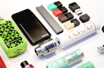 Assorted vaping devices and accessories arranged on white surface including colorful pods, batteries, and cartridges.