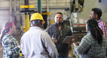 A group of five multiracial mature adults, two women and three men, working in a metal fabrication shop having a meeting on the factory floor. A mixed race Hispanic and Caucasian man is in charge, holding a digital tablet, talking and looking toward an African-American man wearing a hard hat.