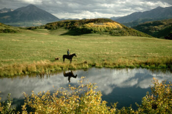 A scenic mountain landscape with a person on horseback and a dog beside a reflective pond. Mountains rise in the background under cloudy skies, while the rider's reflection is mirrored perfectly in the still water, surrounded by autumn foliage.