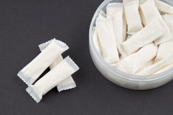 Close-up of white nicotine pouches against a black background, showing both loose pouches and a plastic container filled with them. The pouches have a distinctive rectangular shape with serrated edges.